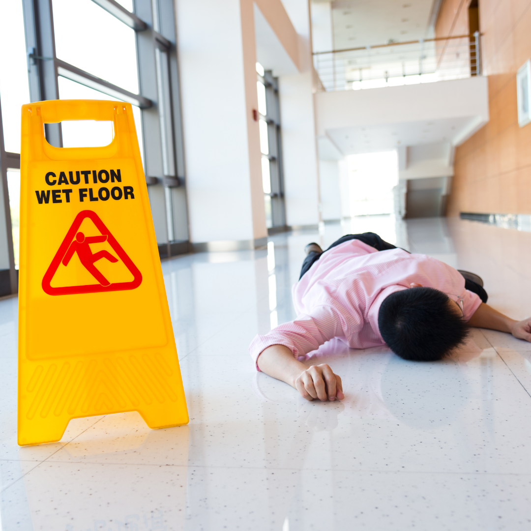 A person lies on the floor next to a yellow "Caution Wet Floor" sign in a bright hallway, suggesting they slipped and fell—an incident where a Jamaica Queens Accident Lawyer could offer helpful legal guidance.